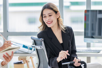 Millennial Asian pretty cheerful successful professional businesswoman in formal suit sitting at working desk helping advising female colleague writing note on notebook in company office workstation.