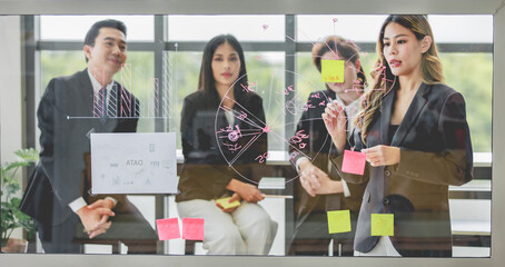Millennial Asian successful professional female businesswoman standing using marker pen writing drawing graph chart data information on glass board presenting to colleagues in office meeting room