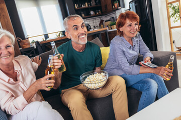 Group of senior friends having popcorn and beer watching tv together during weekend at home