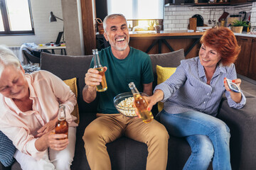 Group of senior friends having popcorn and beer watching tv together during weekend at home