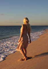 full length portrait of beautiful young woman with long hair wearing flowing dress, standing pose walking away from the camera.  ocean beach background with sunset lighting.
