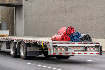 Big rig semi truck running on the road transporting rolled up tarp for covering goods stacked on a...