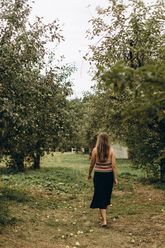 a woman in a black skirt walks barefoot on the trampled grass in an apple orchard. a girl with streaked long hair walks alone in her garden on a cloudy day.