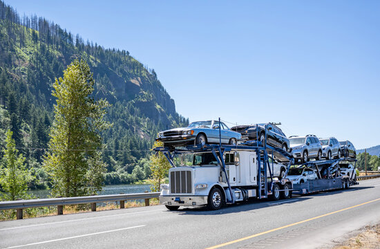 Big rig white classic car hauler semi truck transporting cars on the modular two level semi trailer driving on the one way road along the river
