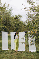 young beautiful brunette woman posing near hung decorative curtains in the garden. a girl in a long green skirt and a white shirt stands barefoot on the grass in an apple orchard