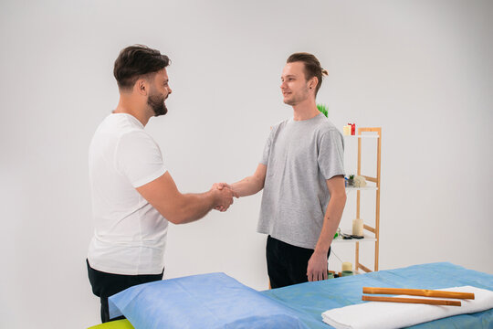 Attractive masseur man shaking hands with his client at the end of procedure at the spa salon in the massage room in front of the camera
