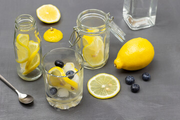 Sliced lemon in glass and in jar. Bottled water. Whole lemon and blueberries on table.