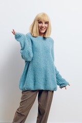 a pleasant, beautiful blonde woman with a short haircut stands on a light background in a long blue sweater in a relaxed pose and happily looks into the camera raising her hand
