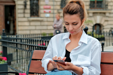 a beautiful girl sits on a bench and holds a phone in her hands. the girl orders food through the phone. the girl is talking on the phone laughing , happy , surprised .