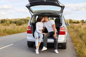 Portrait of loving couple sitting and kissing in their car in the trunk in nature on road, man and woman wearing white shirts traveling together, having romantic journey.