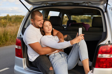 Image of lovely young adult couple sitting together in the car trunk with smart phone and taking selfie or broadcasting livestream, spending time together while traveling.