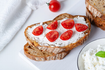 Slices of bread with cottage cheese and cherry tomatoes on wooden cutting board on white background