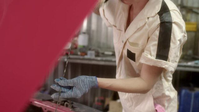 brunette woman mechanic in beige overalls checks oil level in engine of red car in car service. concept of work of car service and maintenance of units in repair shop.