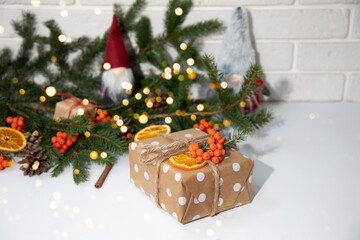 Gift in eco packaging, wrapped in brown paper with white circles and decorated with a dry orange, mountain ash and a fir branch. New Year's gifts in lights on white table, top view