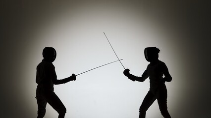Side view of black silhouettes two young female swordswoman with crossed rapiers. Athletes in white uniforms and protective helmet fighting a duel in dark studio with backlight.