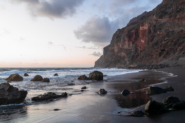 Scenic view during sunset on volcanic black sand beach Playa del Ingles in Valle Gran Rey, La Gomera, Canary Islands, Spain, Europe. Massive cliffs of the La Mercia range. Smooth waves sweeping rocks