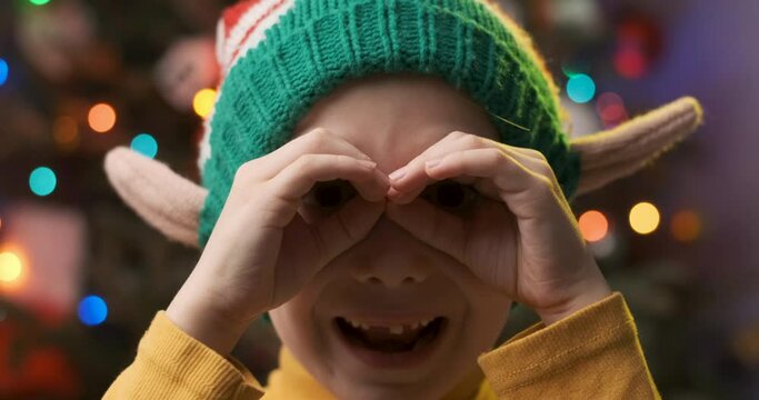 Toothless boy in an elf hat makes binoculars with his hands. Christmas Eve