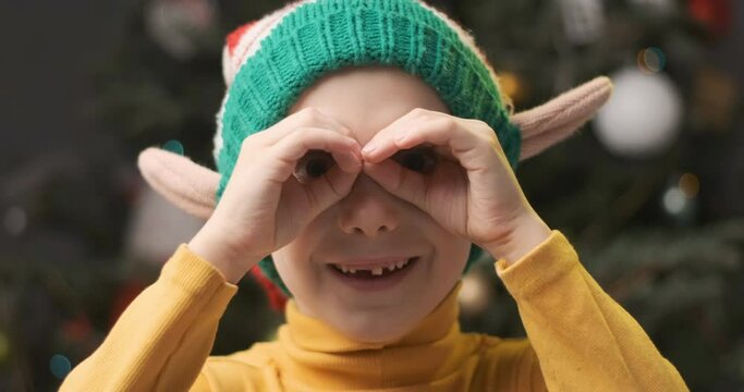 Boy in knitted Christmas elf hat doing gesture like binoculars on Christmas tree background.
