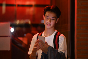 Asian male traveler standing on night city street near showcase and using mobile phone. Night life concept