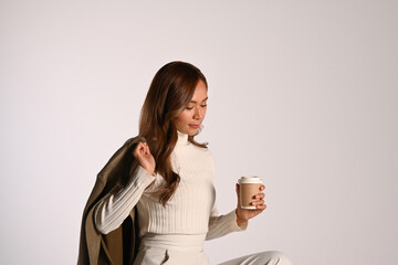 Young beautiful lady in fashionable autumn outfit holding paper cup sitting over light background. Autumn and Winter concept