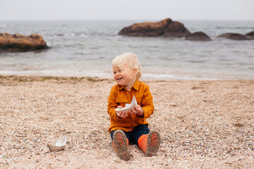 Happy baby playing with paper boat in the beach in autumn time