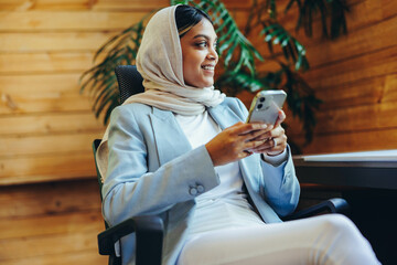 Cheerful Muslim businesswoman using a smartphone in an office - obrazy, fototapety, plakaty