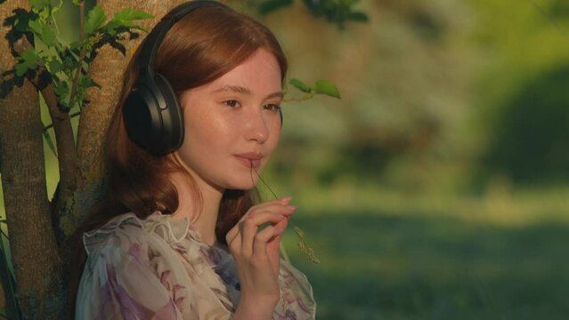 Girl in headphones listen to music, podcast or audiobook, chewing piece of grass outdoors in the country. Handheld video