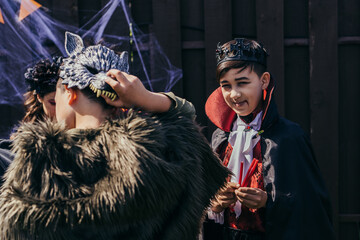 Smiling asian boy in halloween costume holding candies and looking at camera near friends in...