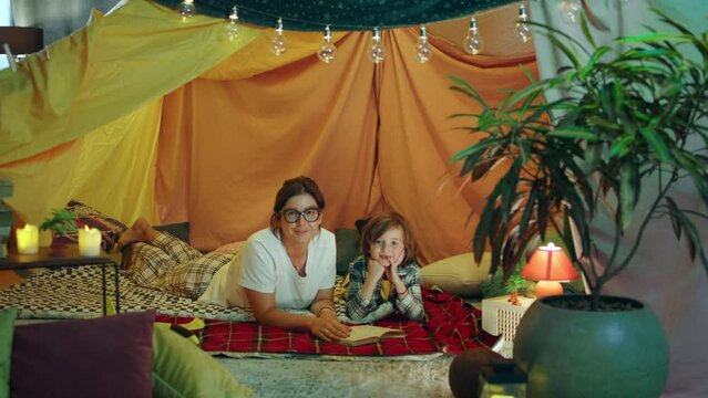 A mother and her charming son are in a very enjoyable looking blanket fort the atmosphere is very relaxing as they are both laying down in it and staring at her camera directly while happily smiling