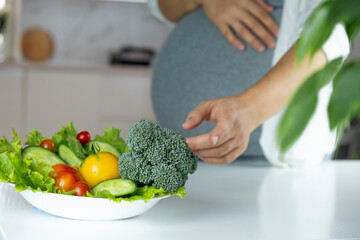 Pregnant woman stretch hand to plate with vegetables and fruits on table. Nutrition and healthy diet during pregnancy. Healthy eating full of vitamins and antioxidants for pregnant woman.