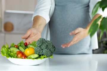 Belly of pregnant woman on background and plate with vegetables and fruits on table. Nutrition and...