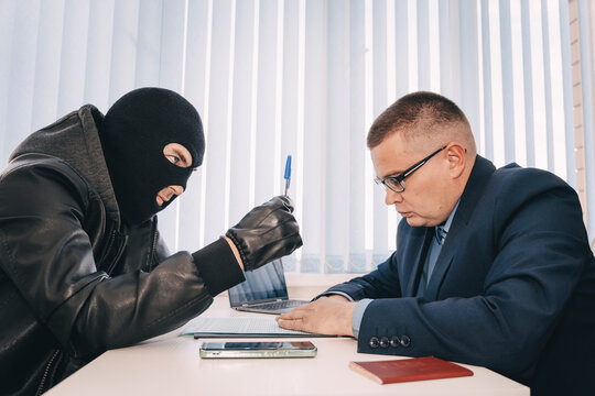 businessman and robbers are sitting at a table. A racketeer in a black balaclava forces to sign a contract. The concept of a raider takeover of the company. dangerous deal