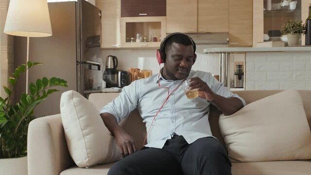 Medium shot of Black man listening to music in headphones while sitting on sofa in his modern minimalist studio apartment