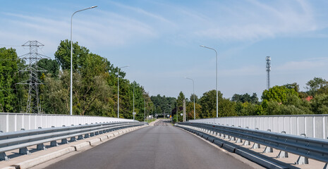 Asfaltowa droga idąca przez most nad rzeką Odrą otoczona barierkami wokół oświetlenie uliczne oraz nieliczne drzewa a na tle błękitne lekko zachmurzone niebo w zachodniej Polsce © FIOMI