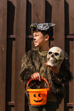 smiling asian boy in werewolf costume looking away while standing with trick or treat bucket and skull