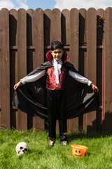 full length of asian boy in vampire costume holding cloak near skull and halloween bucket on grass