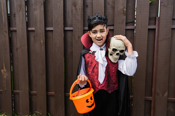 happy asian boy in vampire costume holding skull and trick or treat bucket while looking at camera