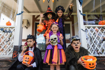 Obraz na płótnie Canvas multiethnic friends in halloween costumes holding buckets of candies and grimacing near decorated cottage