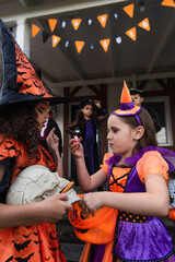 girls in halloween costumes holding skull and bucket with candies near blurred multiethnic friends