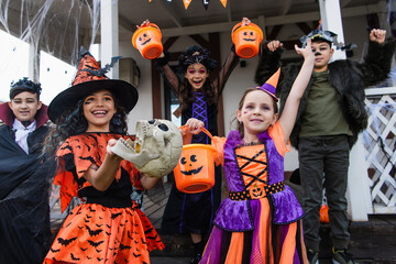 excited multicultural friends in halloween costumes holding trick or treat buckets near house