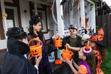 Positive multiethnic kids holding buckets with candies while celebrating halloween in backyard