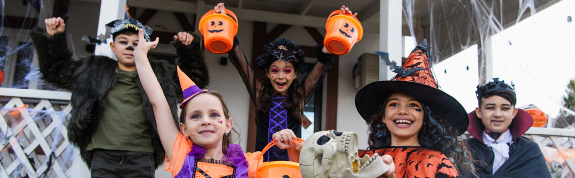 cheerful multiethnic friends in halloween costumes with trick or treat buckets near cottage, banner