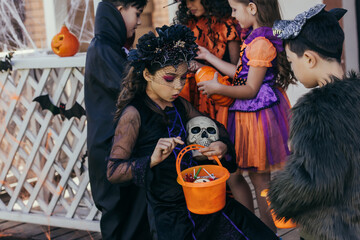 Shocked girl in costume holding bucket with candies near asian friend during halloween celebration outdoors