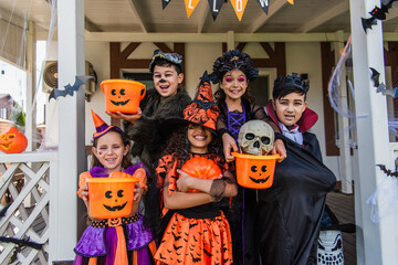Happy multiethnic kids in halloween costumes holding buckets and pumpkin near house outdoors - obrazy, fototapety, plakaty
