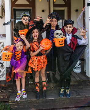 Multiethnic children in halloween costumes holding buckets and grimacing at camera near house
