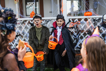 Asian boys grimacing while holding halloween buckets near blurred friends outdoors