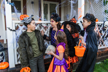 Kids in costumes grimacing near asian friend with bucket near halloween decor in backyard