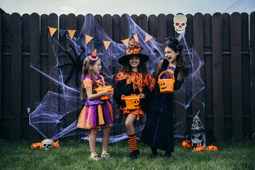Cheerful preteen girls in costumes holding buckets near festive halloween decor on fence outdoors