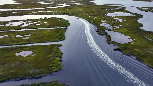 An aerial view over the salt marshes in Hempstead, NY on a cloudy day. The drone camera dolly in tilt down chasing a small boat speeding by in the calm waters, maneuvering through the channels.