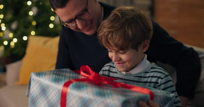 Curious child enjoys the surprise of a Christmas gift, unties the bow shakes the box with interest, dad looks at his son with joy and pride.
